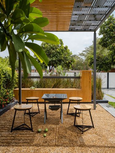 Outdoor pergola seating area with wooden stools, fire pit table, and gravel flooring surrounded by greenery