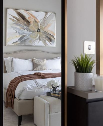 Beige upholstered bed with white linens and rust throw seen through a mirrored doorway, plant and console beyond