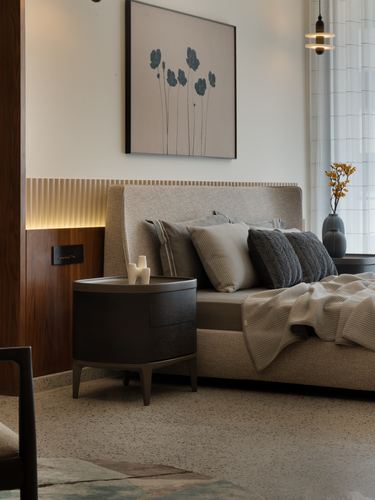 Bedroom with upholstered bed, ribbed wood accent wall with cove lighting, round dark bedside table, terrazzo floor.