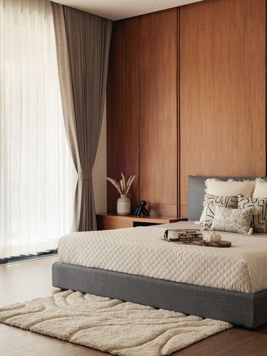 Bedroom with wood-paneled wall, grey upholstered bed, sheer curtains, and textured cream rug