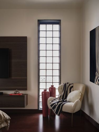 Bedroom corner with tall grid window, cream wingback chair, and dark red ceramic vessels