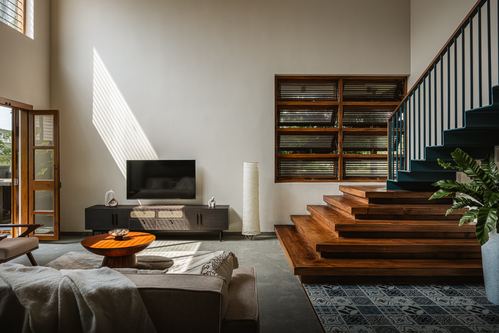 Double-height living room with wooden stair steps, louvered window, sofa and sunlight streaks