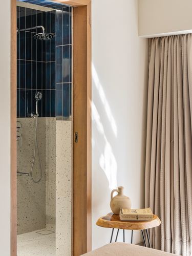 Doorway view into a blue-tiled shower beside a sunlit bedroom corner with curtain and stool