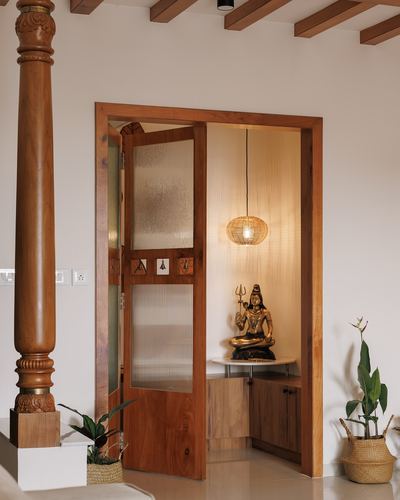 Traditional pooja room with carved wooden pillar, fluted glass doors, and lit brass deity niche