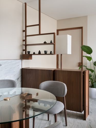 Dining nook with wooden shelving unit, glass table, grey chairs, and cabinet against beige walls