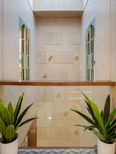 Double-height stairwell with arched windows, fluted stone wall panel, brass bird details, and potted plants behind glass railing.