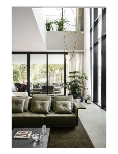 Double-height living room with olive leather sofa, looping brass pendant light, and full-height black-framed glazing onto garden