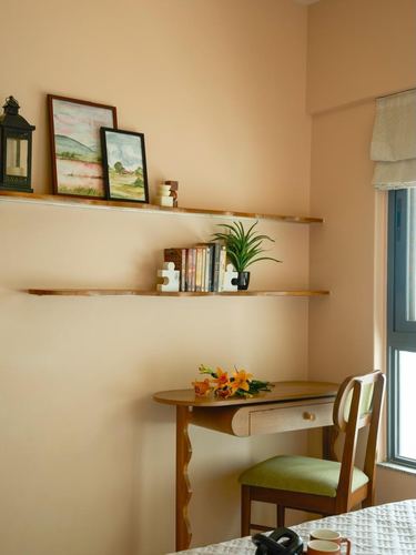 Peach-walled study nook with floating shelves, framed art, and a small wooden desk with chair