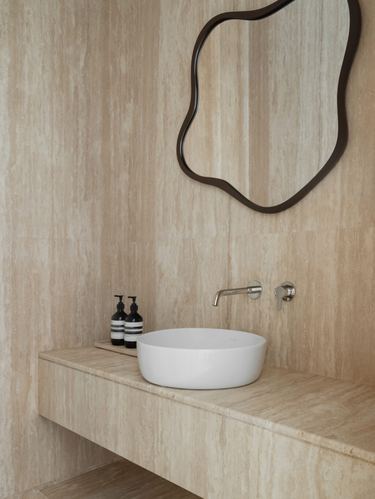 Travertine bathroom with round vessel sink, wall-mounted faucet, and organic-shaped mirror
