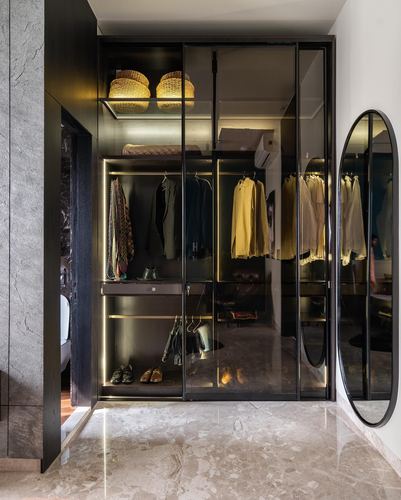 Dark glass-fronted wardrobe with backlit shelves, hanging clothes, and an oval mirror beside marble flooring