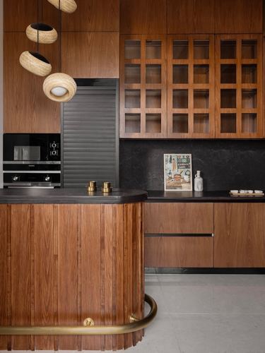 Warm wood kitchen with curved fluted island, brass tap, and rattan pendant lights over dark stone counters