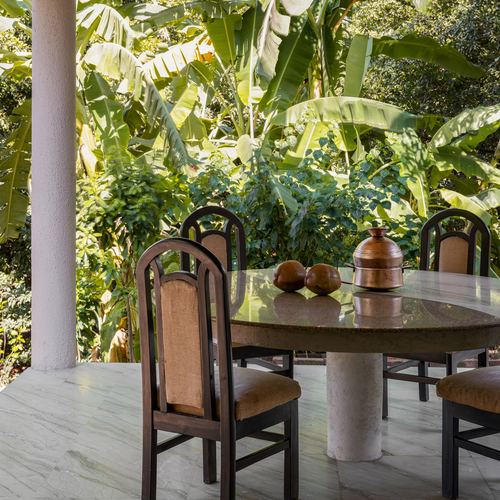 Round stone dining table with cane-back chairs on a marble veranda facing dense banana leaf foliage