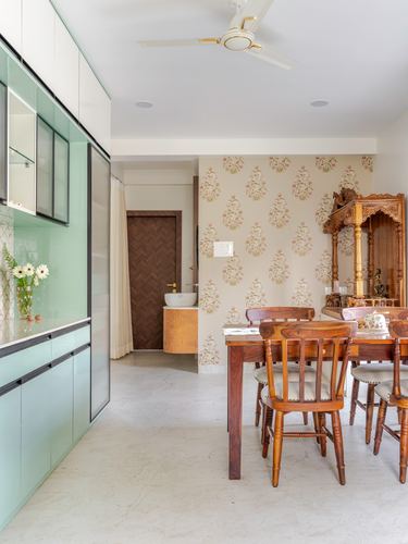 Dining area with wooden table and chairs beside floral wallpaper and carved wooden mandir