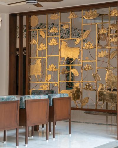 Gold brass screen with lotus and cow motifs behind wooden dining chairs on marble floor