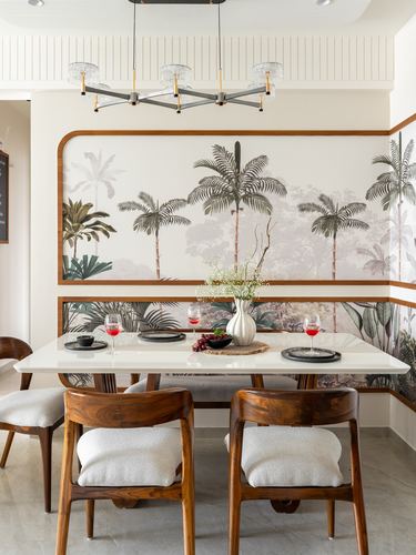 Dining table with wooden chairs set before a curved banquette and palm mural, chandelier above