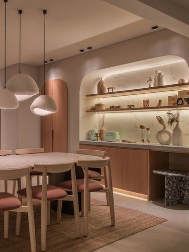 Warm-toned dining nook with round wood table, pink chairs, and backlit arched shelving display