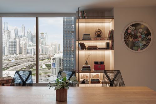Dining table with wire chairs facing a backlit shelving unit and floor-to-ceiling Dubai skyline window