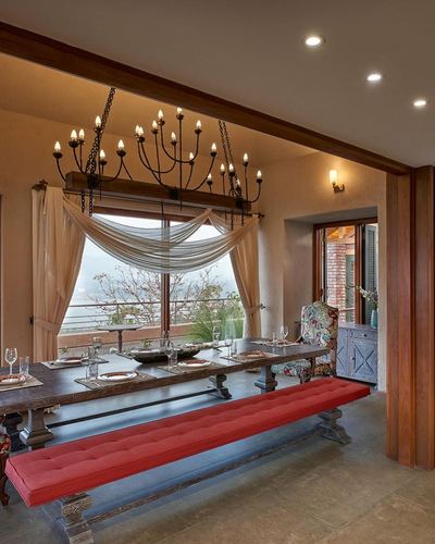 Rustic dining room with iron chandelier over long wood table and red tufted bench, valley view through window