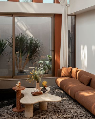 Sunlit living room corner with curved brown sofa, travertine coffee table, and glazed doors overlooking greenery