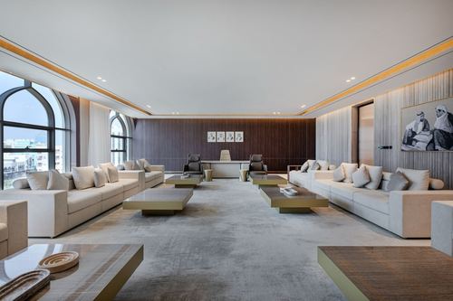 Symmetrical majlis-style lounge with cream sofas, arched windows, and a wood-paneled far wall leading to a desk