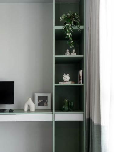 Sage green open shelving with plants and figurines beside a white desk and curtain wall.