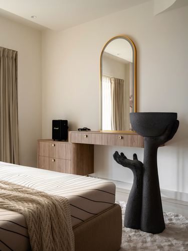 Bedroom corner with wood vanity, arched brass-framed mirror, and black sculptural hand stool.