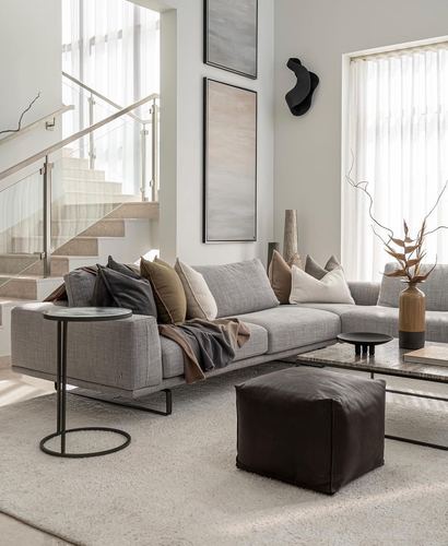 Neutral toned living room with grey sectional sofa, glass staircase, and leather pouf