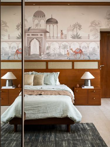 Bed with wood headboard against a hand-illustrated palace mural wallpaper, flanked by matching lamps