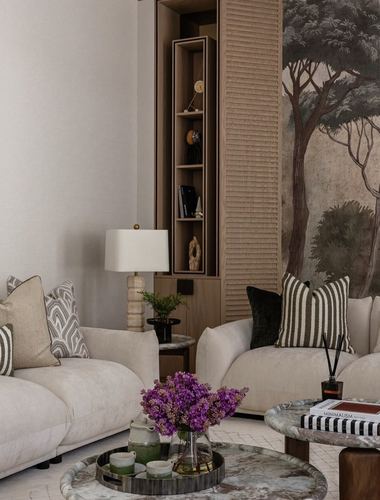 Beige living room corner with two sofas, patterned cushions, and a tree-mural wall behind wooden shelving