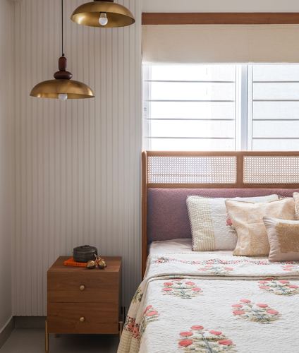 Bedroom corner with cane headboard, mauve upholstery, floral quilt, and two brass dome pendant lights over a wooden nightstand.
