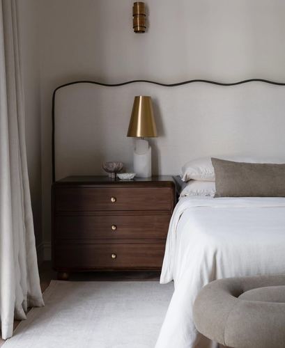 Bedroom corner with wavy linen headboard, dark wood dresser, and brass lamp and sconce