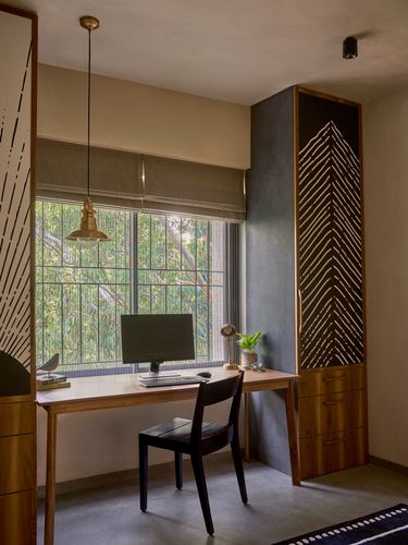 Study nook with wooden desk, black chair, brass pendant light, and carved wood cabinet beside a large window with greenery.
