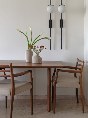 Wooden dining table with two vases of flowers, flanked by cane-detailed armchairs and black wall sconces
