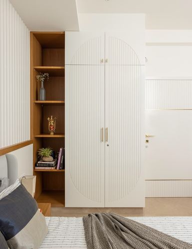 Arched fluted white wardrobe beside open wooden shelving niche with décor in bedroom