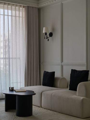 Beige curved sofa with black cushions beside a dark organic coffee table near sheer curtains and panelled wall