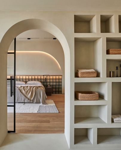 Arched plaster doorway beside a niche shelving wall reveals a bedroom with tiled headboard and cove lighting