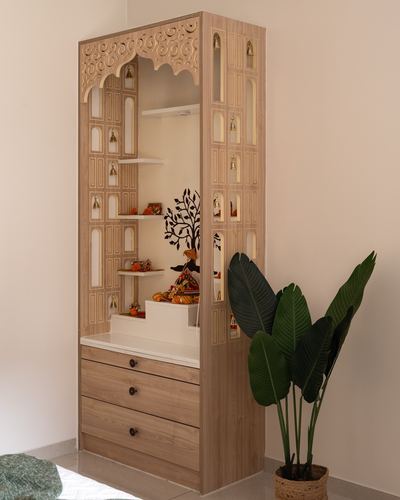 Wooden pooja unit with carved arch and jali panels in a corner, with idols and drawers below