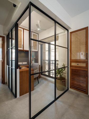 Glass and black steel partition encloses a study nook with cane cabinetry and stone flooring.