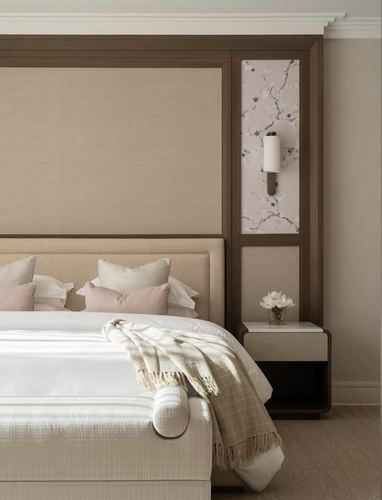 Beige upholstered bed with wood trim, blush pillows, and marble-top nightstand under a floral wallpaper niche