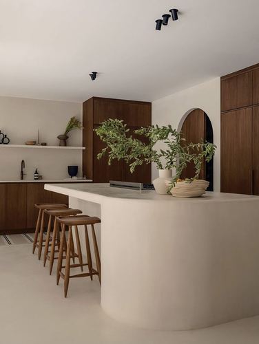 Curved plaster kitchen island with wood stools and dark timber cabinetry, arched niche behind