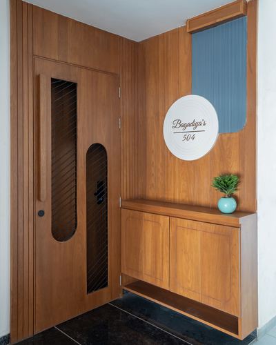 Wooden entry door with grille inserts beside a floating console and circular nameplate reading Bagadiya's 504