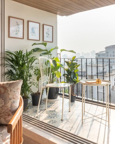 Sunlit balcony with patterned tile floor, potted plants, wire chair and round side tables overlooking Mumbai rooftops