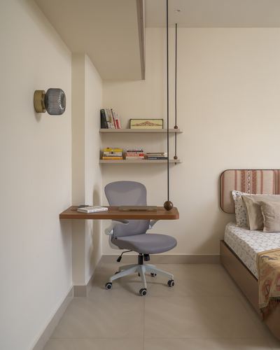 Small wall-mounted wooden desk with grey office chair beside floating bookshelves and a bed