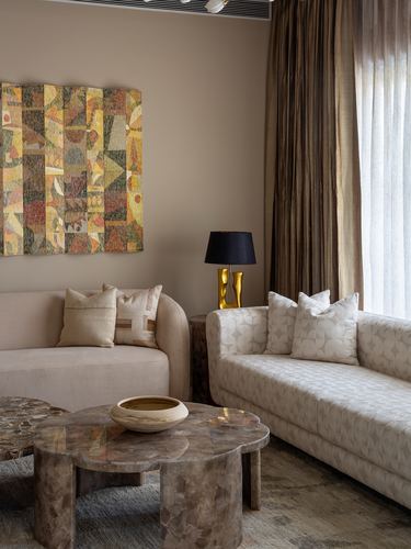Beige living room corner with curved sofas, stone coffee table, geometric wall art, and gold table lamp