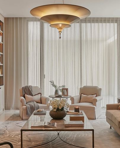 Cream armchairs flank a glass coffee table beneath a tiered brass pendant and sheer floor-to-ceiling curtains