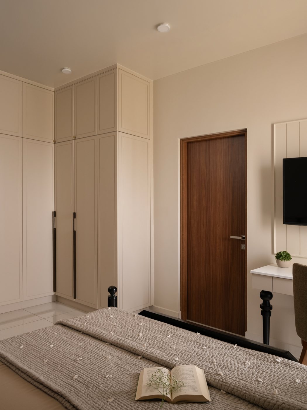 Beige bedroom with tall wardrobe, walnut door, and bed with knit throw and open book