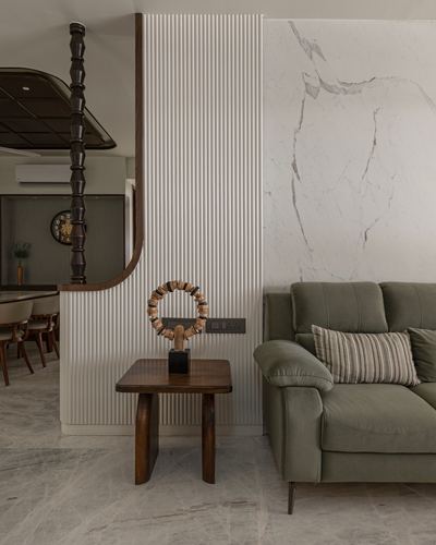 Living room corner with green sofa, wooden side table, sculptural object, and marble-clad wall beside a fluted partition.