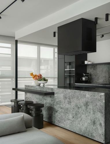 Grey stone kitchen island with black cabinetry, black stools, and sheer-curtained window in a minimalist apartment