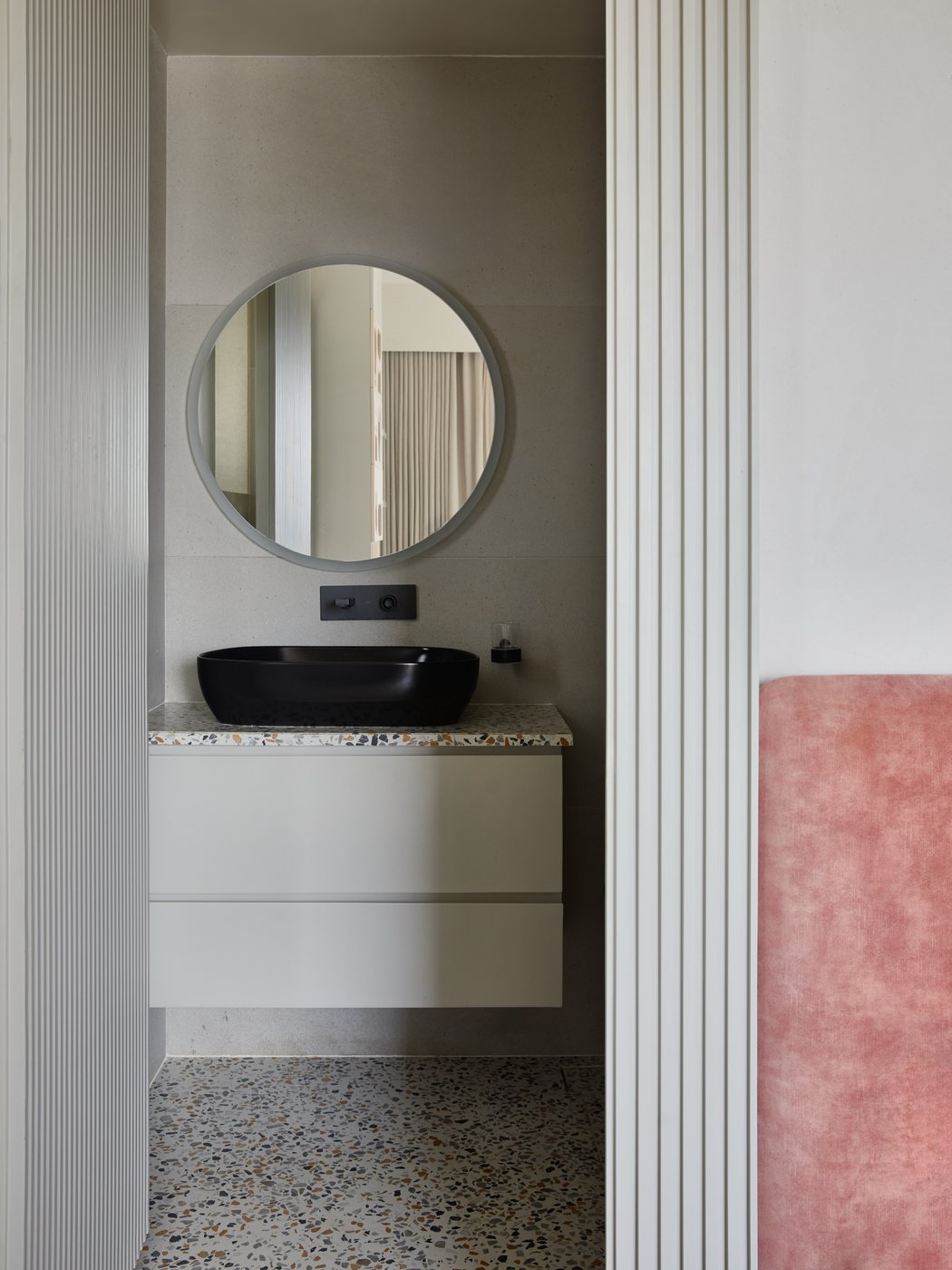 Round mirror above black vessel sink on terrazzo-topped vanity, framed by fluted door panels