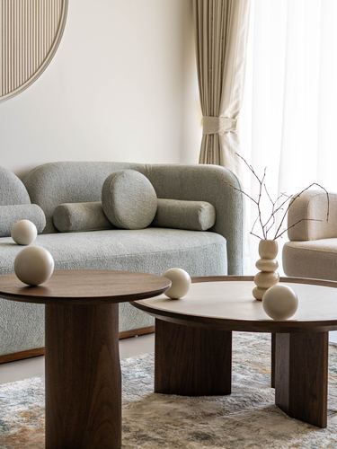 Curved taupe sofa with bolster cushions behind two round wooden pedestal coffee tables